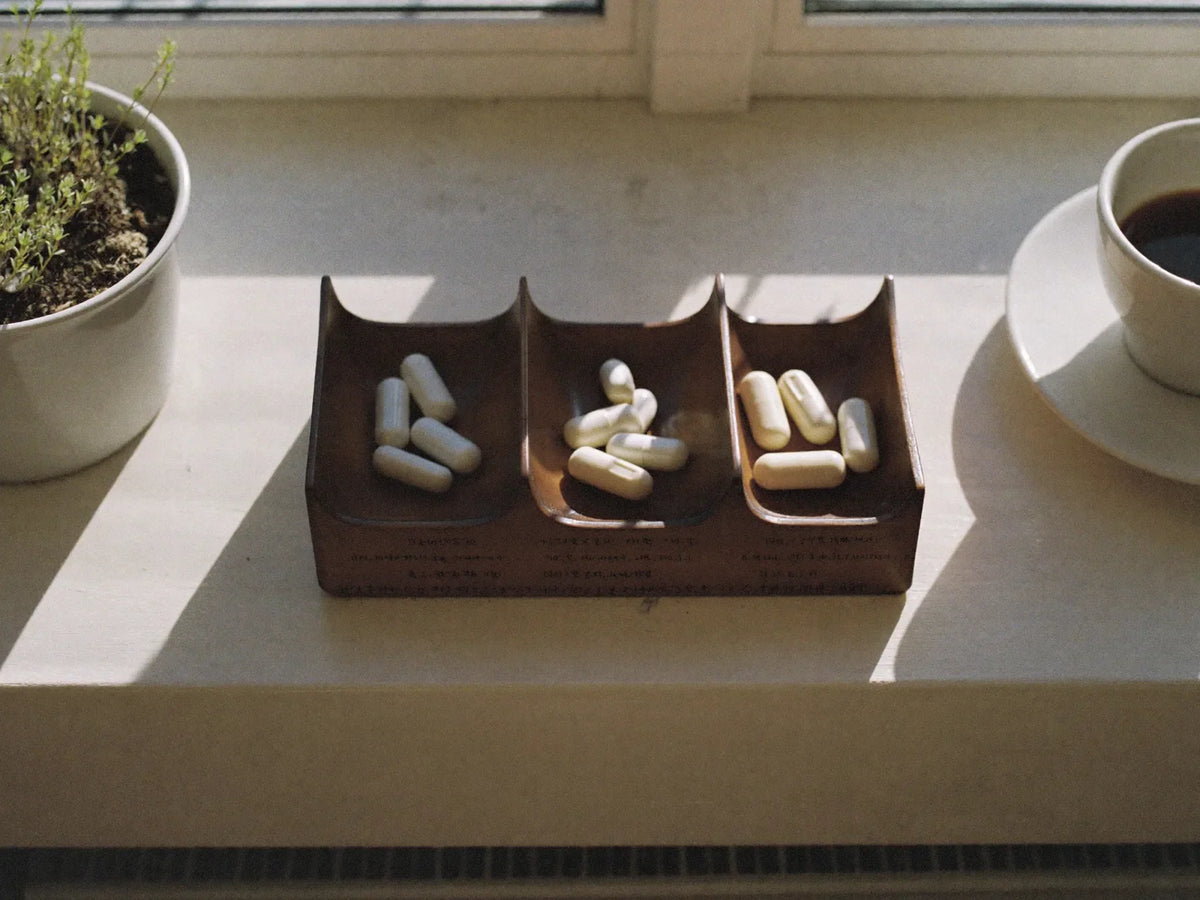 Wooden pill organiser with capsules on a sunlit windowsill beside a coffee cup