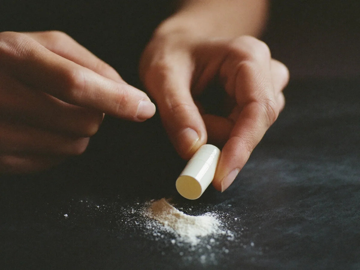 Hands opening a capsule so white powder spills onto a dark surface