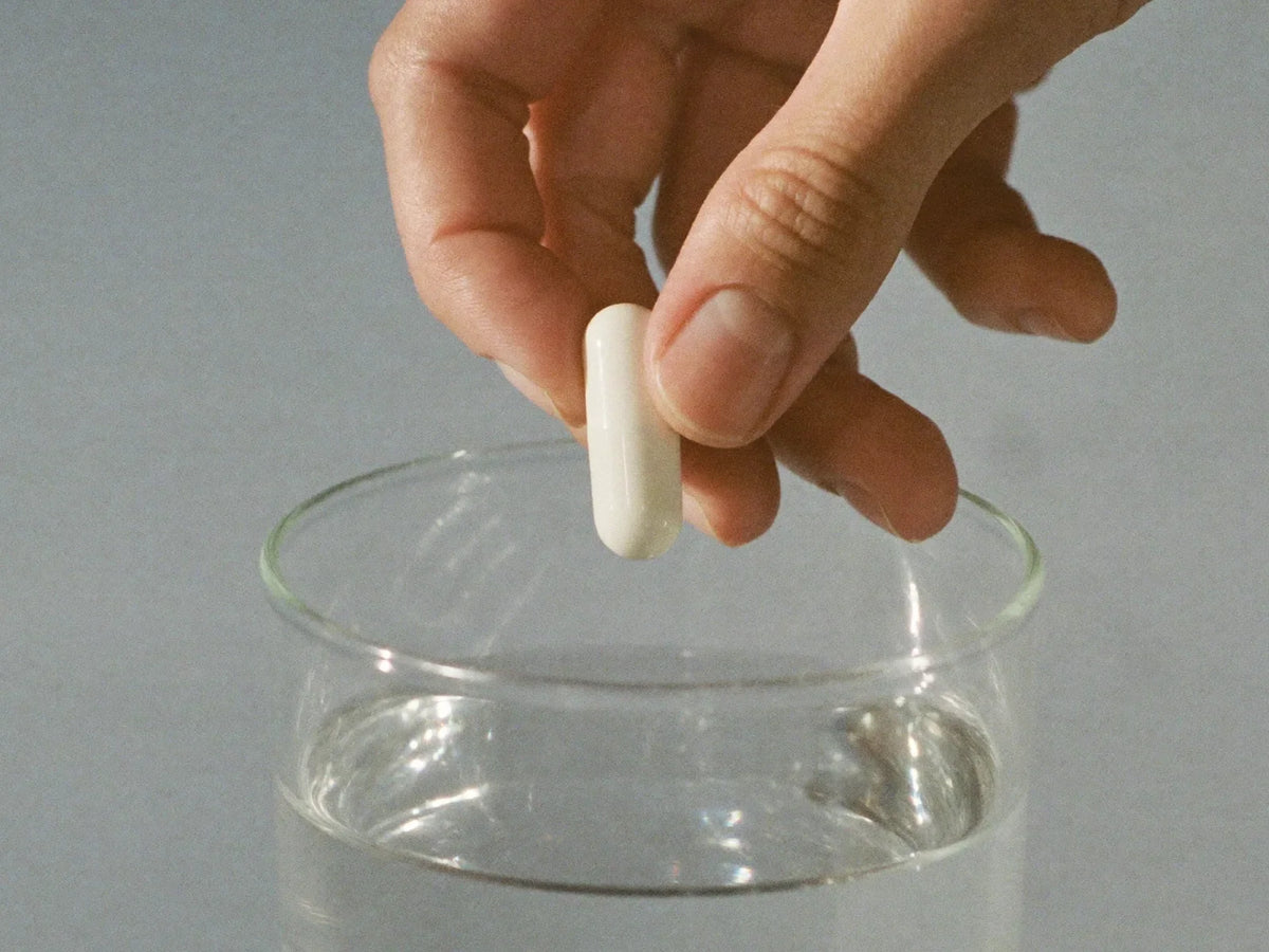 Hand holding a single white capsule above a glass of water