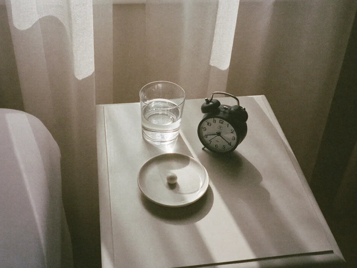 Bedside table in morning light with a glass of water, a capsule in a dish and an alarm clock