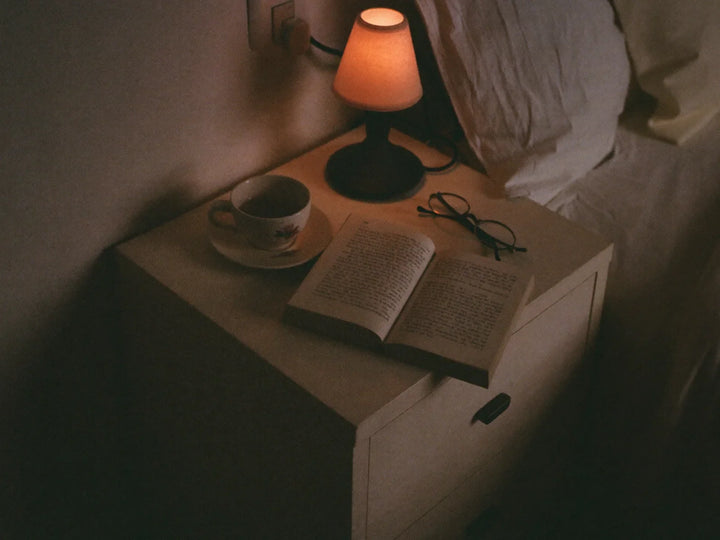 Nightstand at dusk with herbal tea, an open book, reading glasses and a lamp